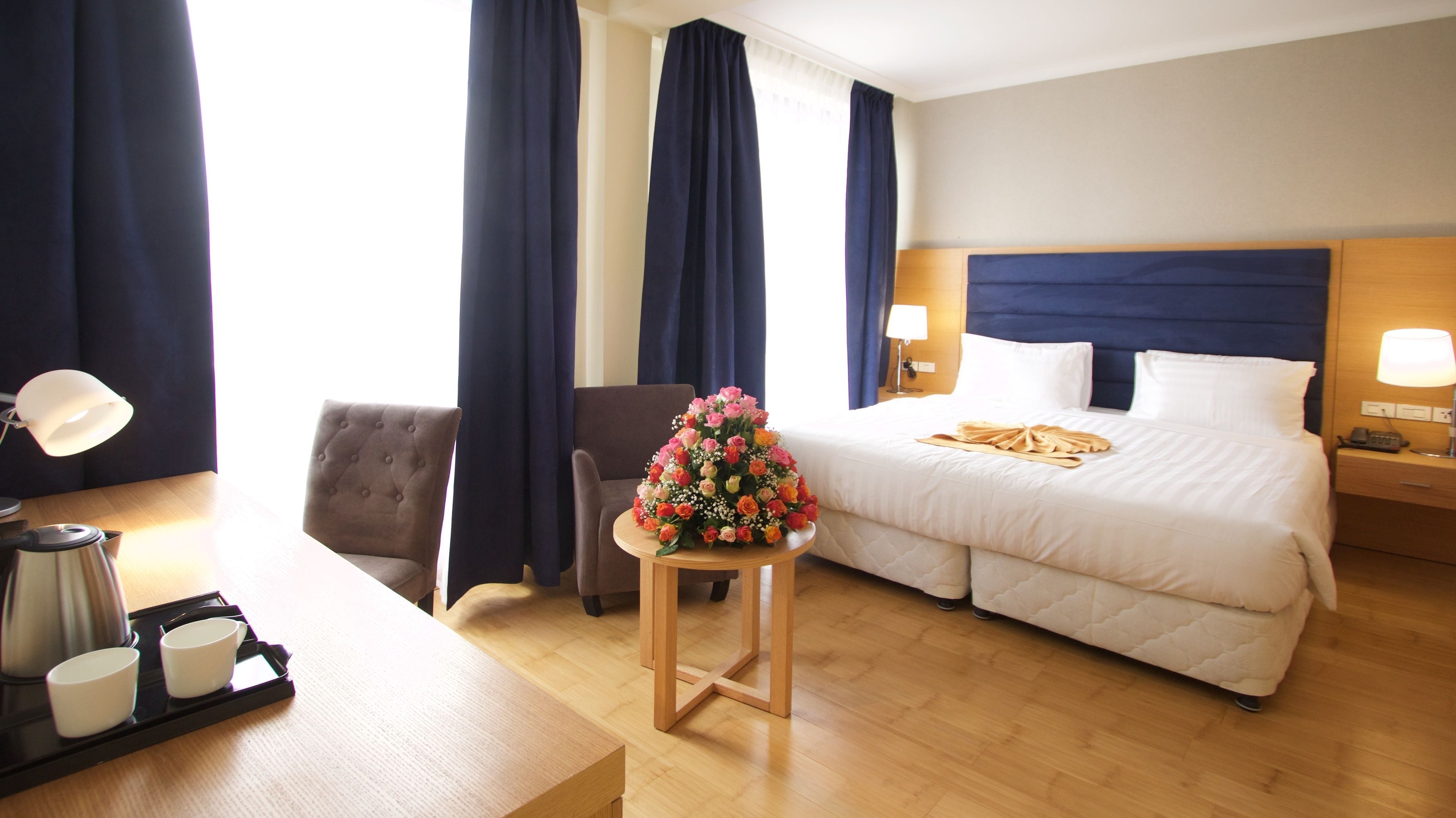 Habitación de hotel con cama, lámparas de noche, escritorio con lámpara, silla, cafetera y arreglo floral.