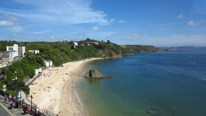 A coastal beach with a clear view of the sea, a sandy shore, and a few buildings in the background. - Sea View Apartment Tenby, Pembrokeshire, Wales. Near Beach, Restaurants & Shops (Tenby)