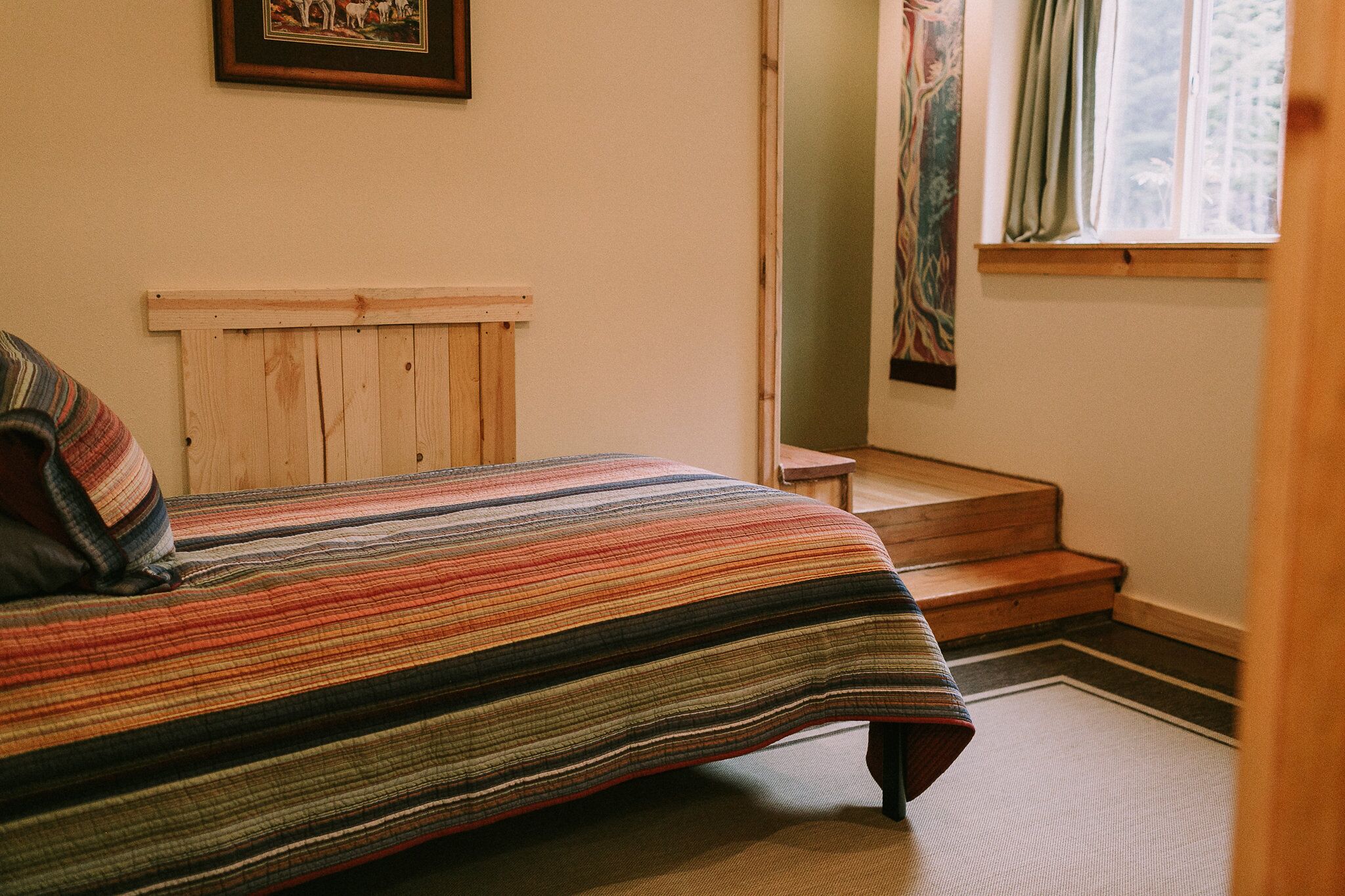 Een slaapkamer met een bed, een houten hoofdbord, een raam met gordijnen en een deur die leidt naar een trap.