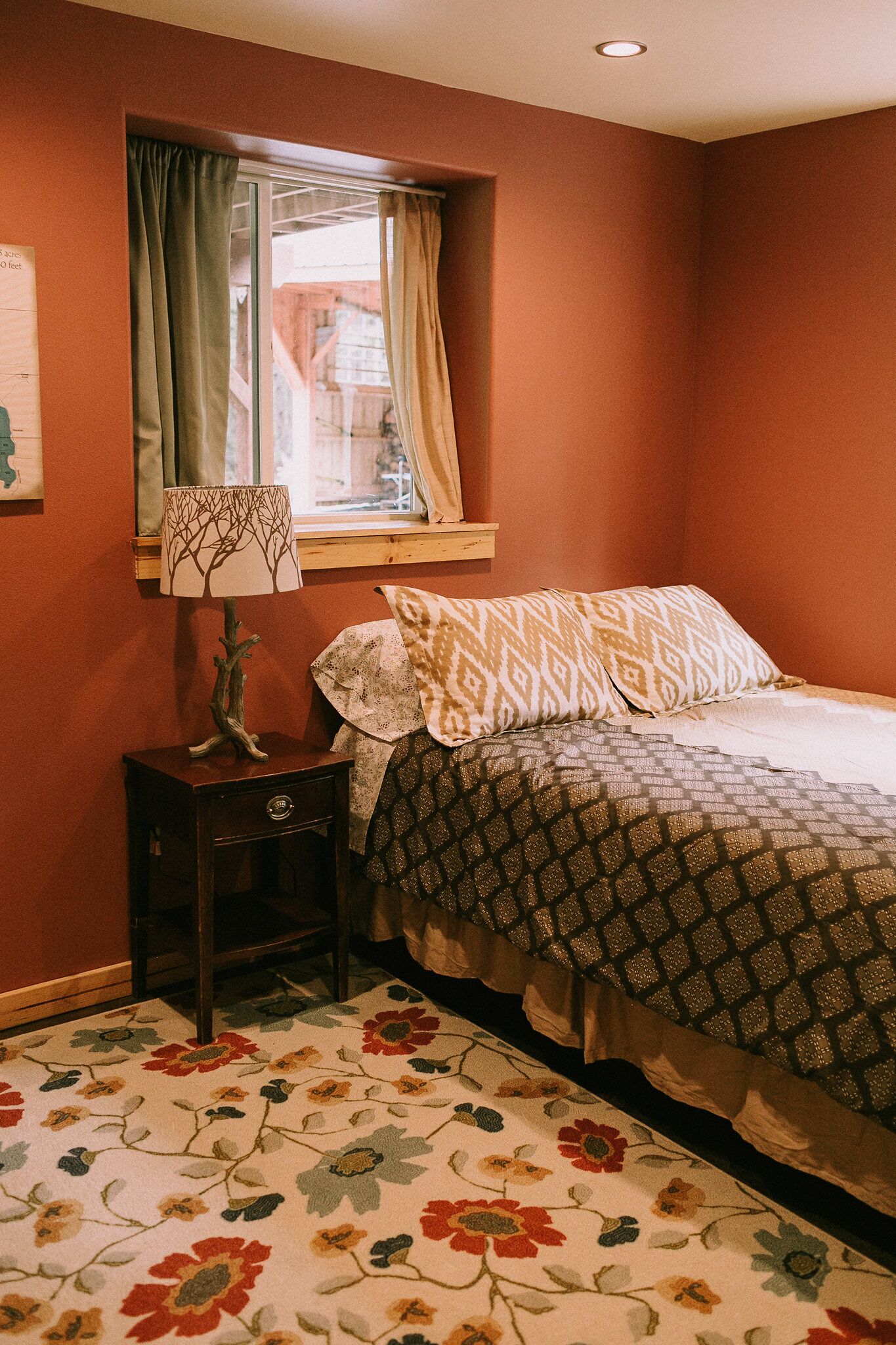 A bedroom with a bed, a nightstand, a lamp, a window with curtains, and a floral carpet.