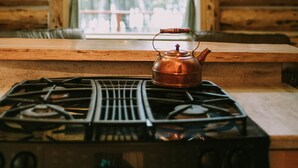 Ein rustikales Blockhaus-Interieur mit Holzvertäfelung, einem Fenster mit Blick auf Bäume und einem Kupferkessel auf dem Herd.