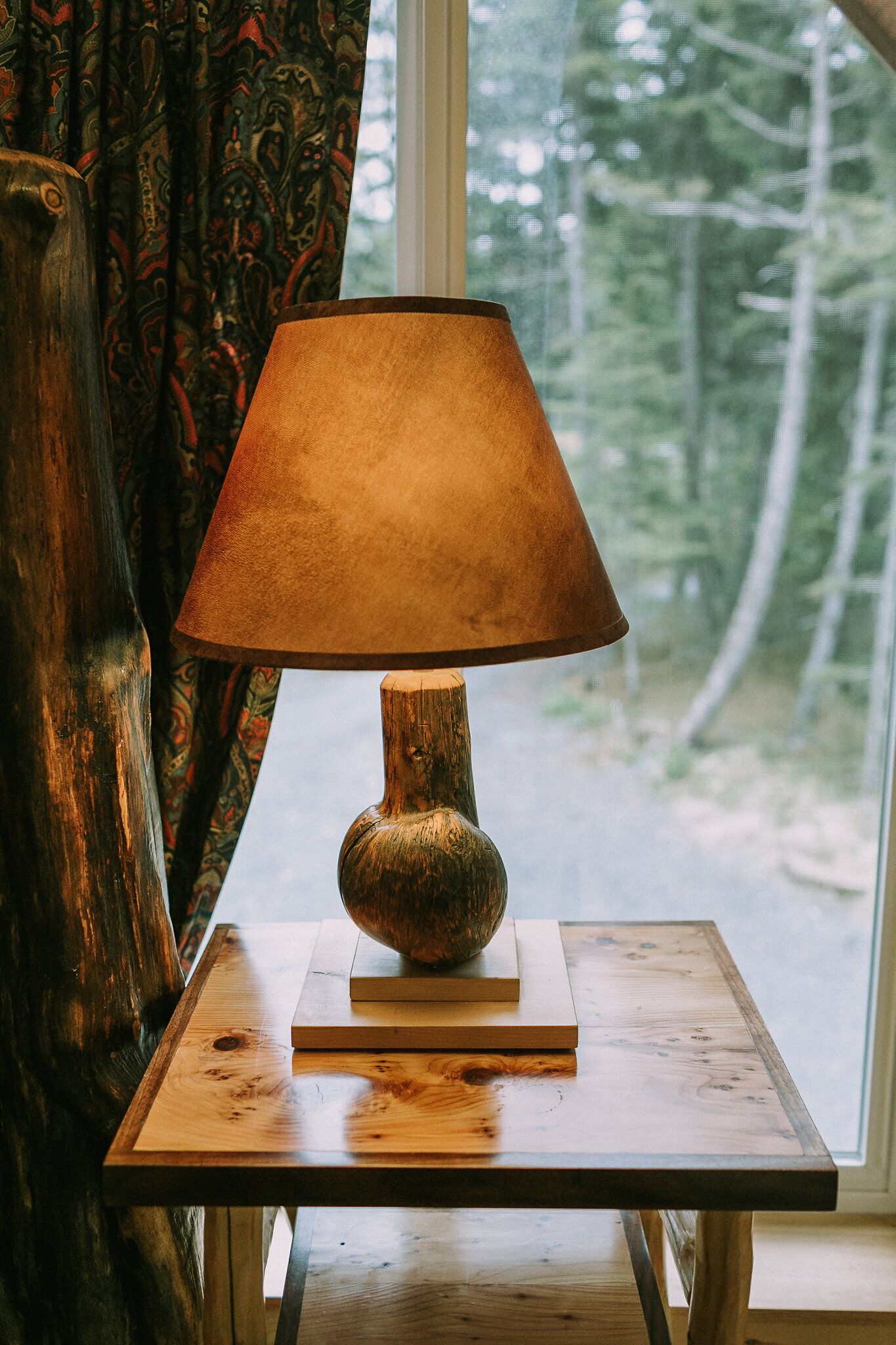 A table with a lamp, a window with a view of trees, and a patterned curtain.