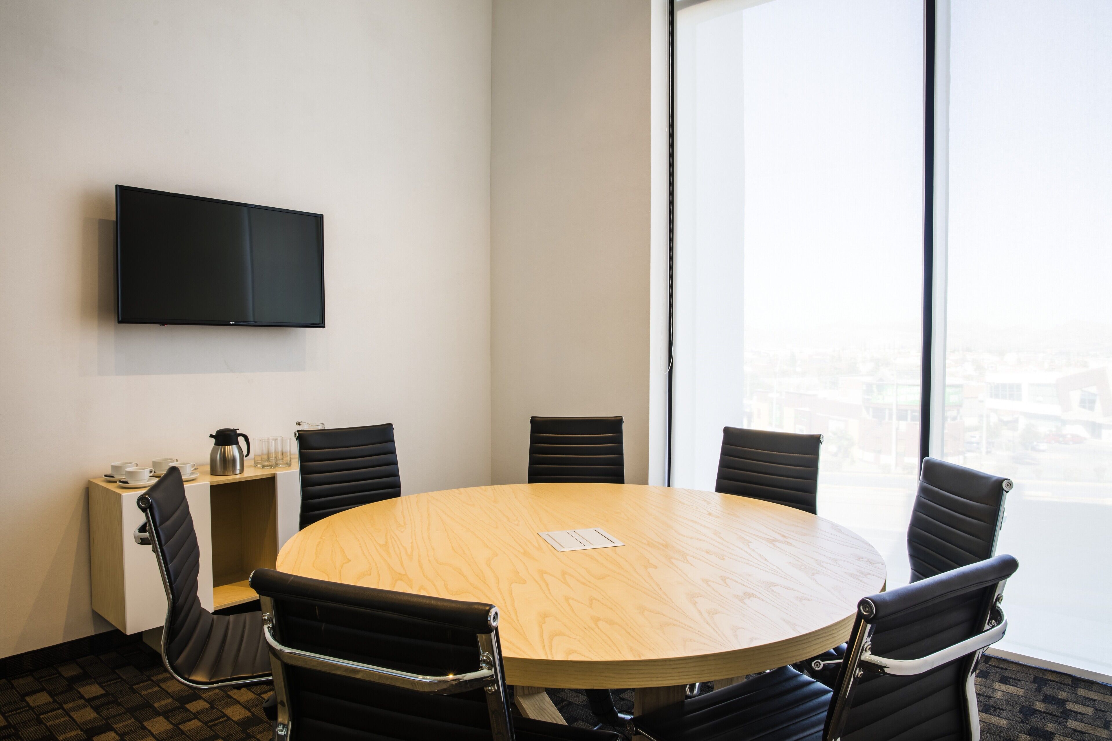 A conference room with a round table, six chairs, a flat-screen TV mounted on the wall, and a window offering a city view.