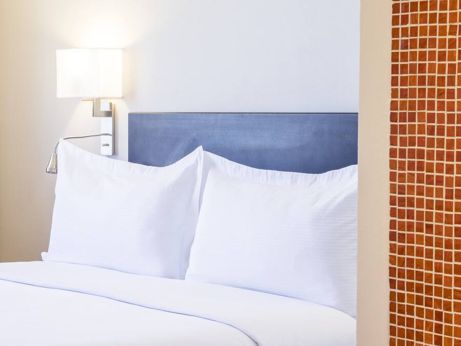 Ein Hotelzimmer mit einem ordentlich bezogenen Bett, einer Nachttischlampe und einer gefliesten Wand.