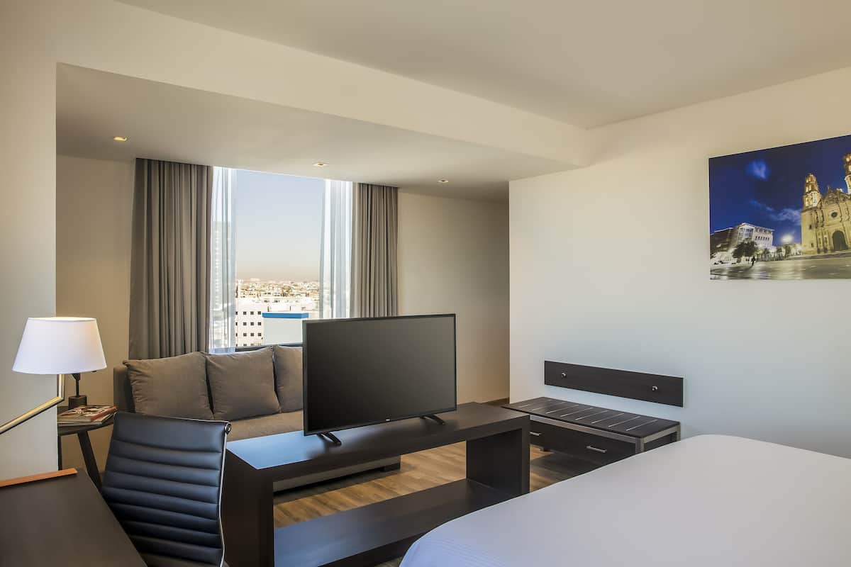 Une chambre d’hôtel moderne dotée d’un grand lit, d’un téléviseur à écran plat, d’un bureau avec une lampe et offrant une vue sur le paysage urbain grâce à de grandes fenêtres.