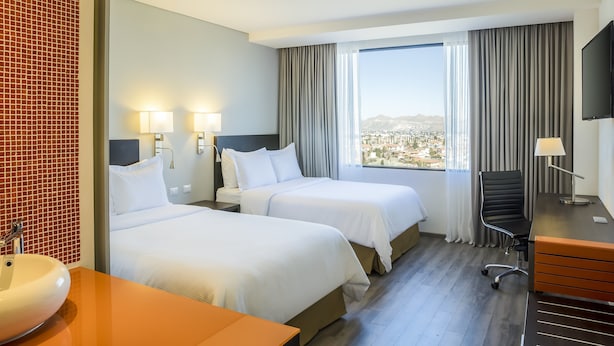 Une chambre d’hôtel avec deux lits, un bureau, une chaise, une télévision et une grande fenêtre donnant sur la ville.