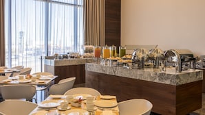 Un espace pour le petit-déjeuner à l’hôtel, avec un buffet, des tables, des chaises et une vue sur la ville grâce à de grandes fenêtres.
