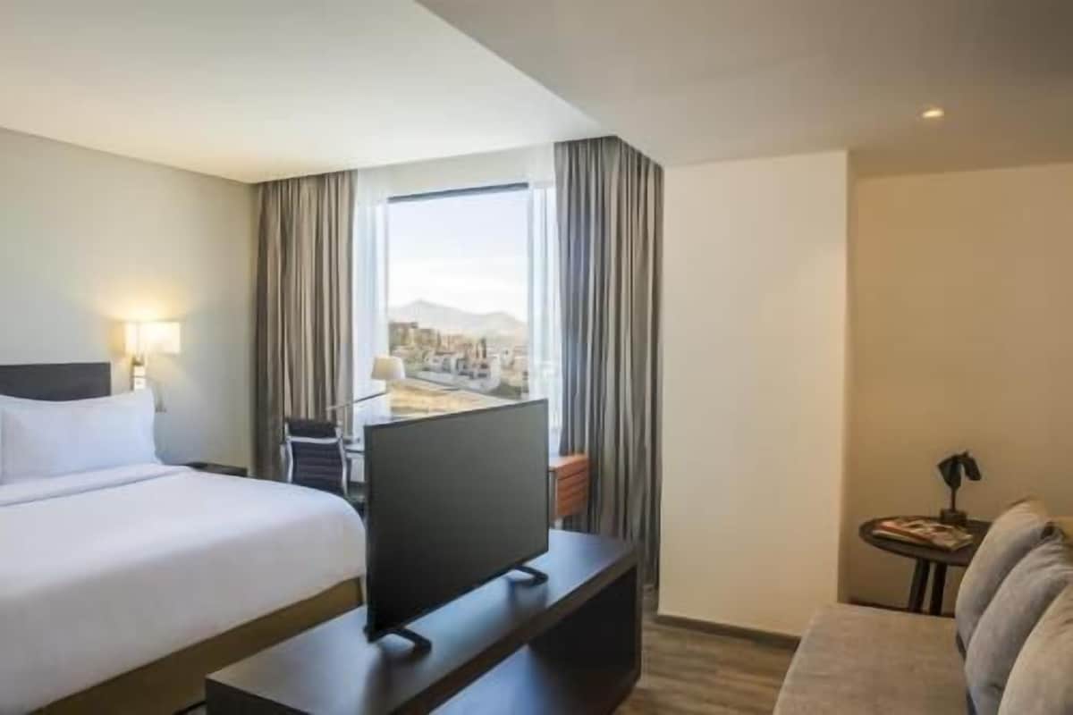 Ein Hotelzimmer mit Bett, Fernseher auf einem Tisch, einem kleinen Beistelltisch und Blick auf eine Stadtlandschaft durch das Fenster.