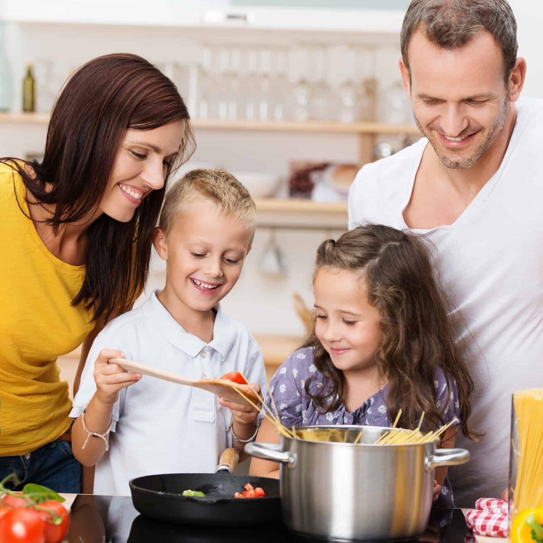 A family cooking together in a kitchen with a pot of spaghetti, a frying pan, and fresh tomatoes on the counter.