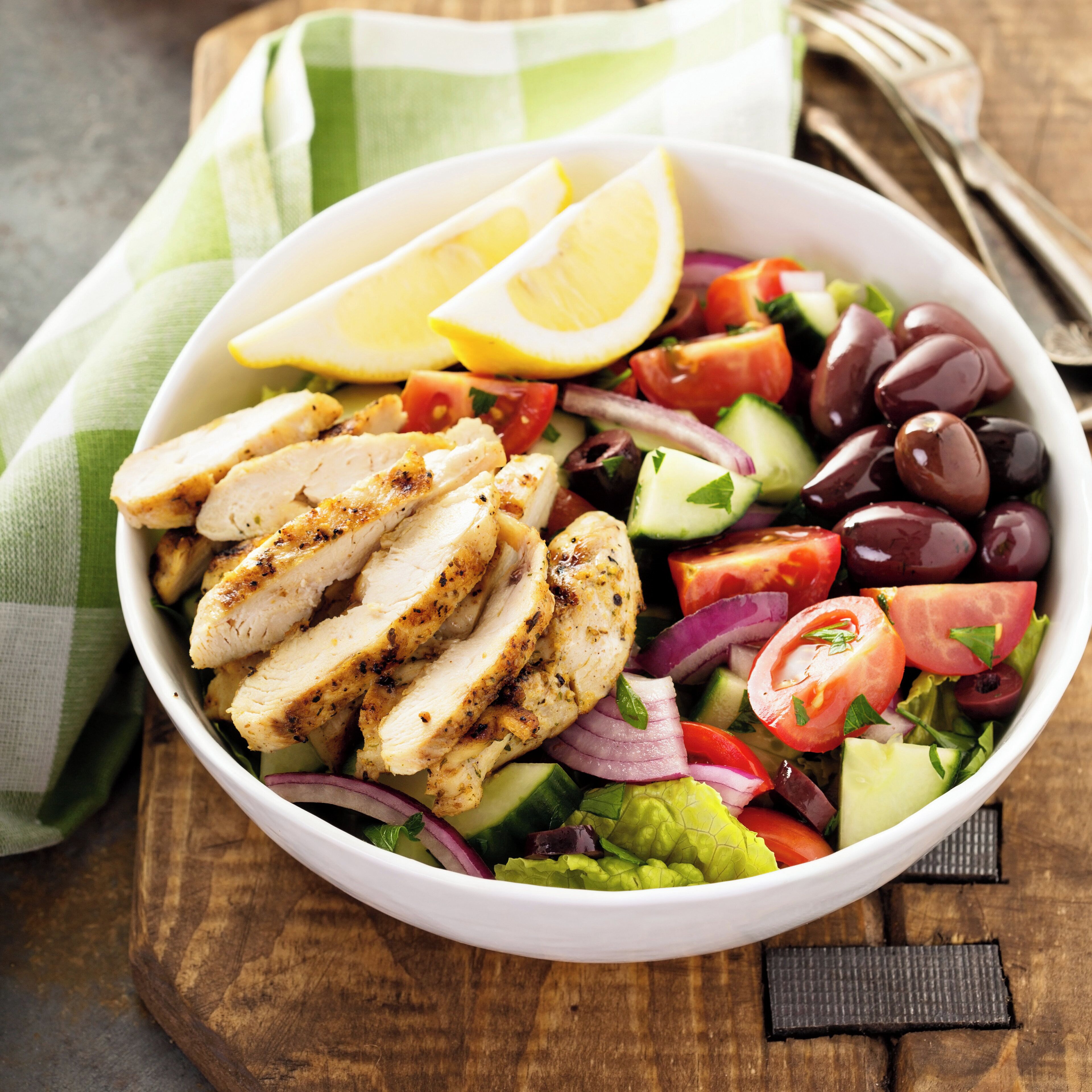 A bowl of salad with chicken, tomatoes, cucumbers, olives, and lemon slices.