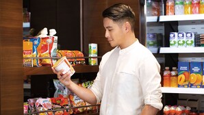Eine Person in einem weißen Hemd hält eine Suppendose in einem Gang eines Supermarktes, wo verschiedene verpackte Lebensmittel und Getränke in Regalen zu sehen sind.