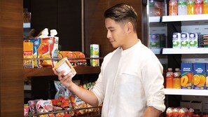 A person in a white shirt holding a can of soup in a grocery store aisle with various packaged foods and beverages on shelves. - Staybridge Suites Doha Lusail by IHG (Doha)