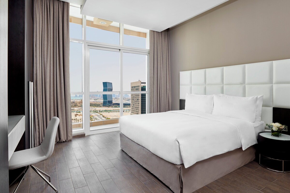 A hotel room with a large bed, a desk, a chair, and a view of the cityscape.