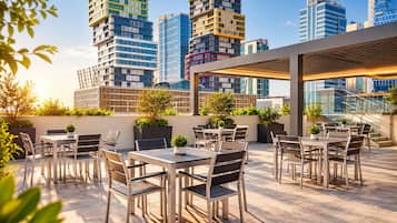 Un espacio al aire libre con mesas y sillas, rodeado de vegetación y vistas a un paisaje urbano con edificios modernos.