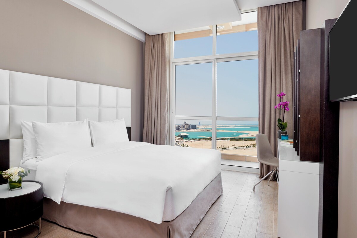 A hotel room with a large bed, a TV, a desk, and a view of the beach.