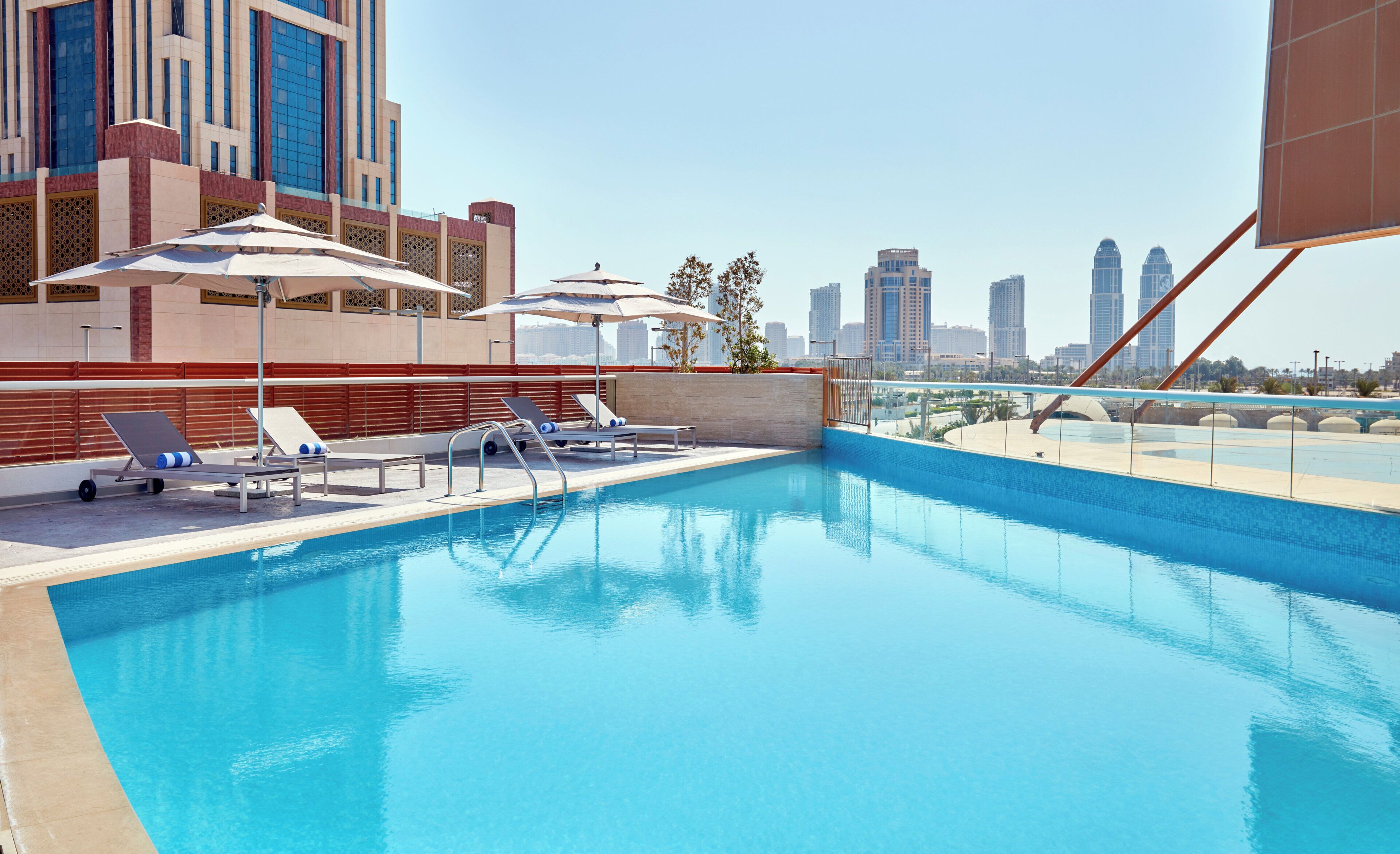 Una piscina en la azotea con sillones de descanso y sombrillas, rodeada de edificios altos.