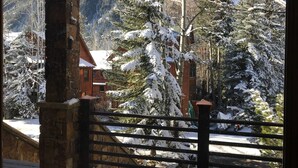 Une cabane en bois avec une entrée en pierre, entourée d’arbres couverts de neige et dominée par un paysage montagneux.