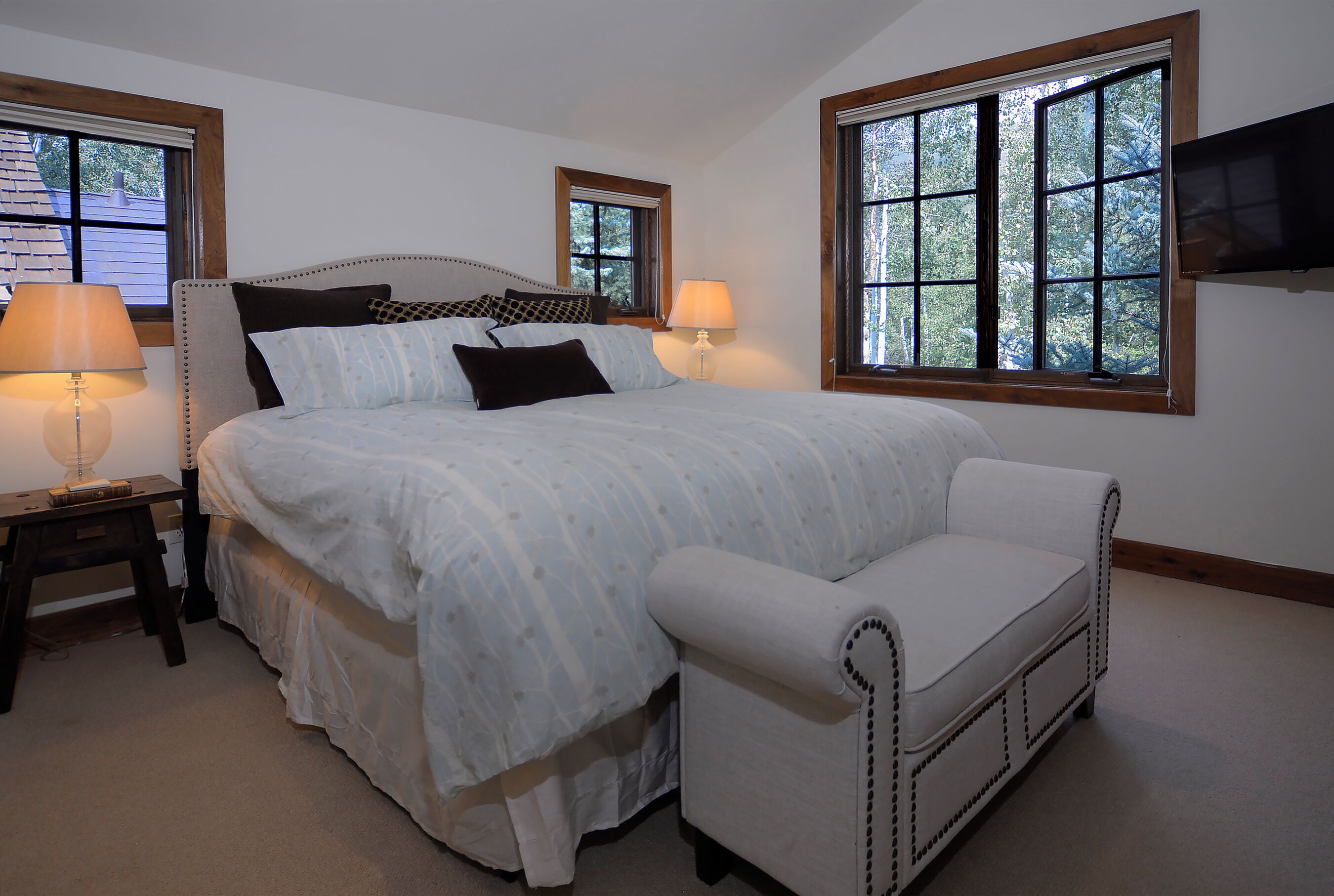 A bedroom with a large bed, two bedside tables with lamps, a white armchair, and a TV mounted on the wall.