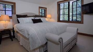 A bedroom with a large bed, two bedside tables with lamps, a white armchair, and a TV mounted on the wall.