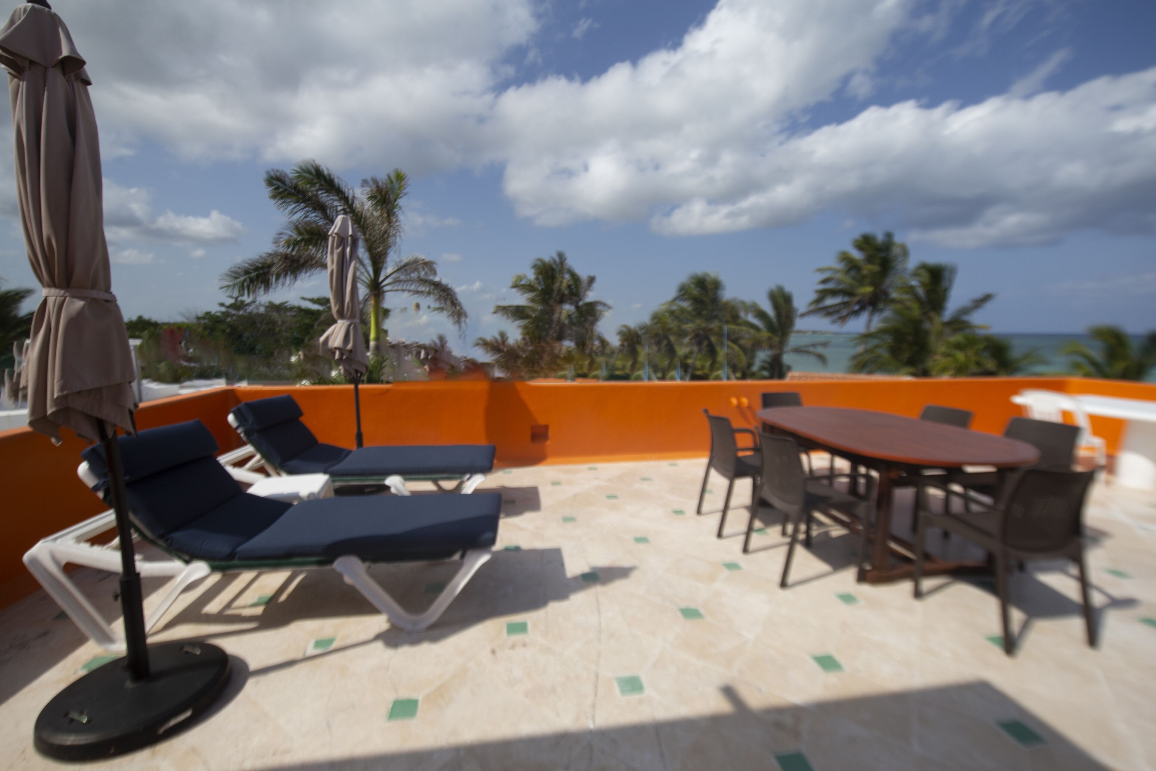A rooftop terrace with lounge chairs, a table, and chairs, overlooking palm trees and the ocean.