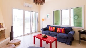 Ein Wohnzimmer mit einem blauen Sofa, einem roten Tisch, Korbnäpfchen und einem großen Fenster mit grünen Fensterläden.