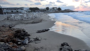 Ein Strand mit einer Reihe weißer Liegestühle und Sonnenschirme, felsiger Küste und einem Gebäude im Hintergrund.