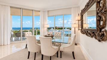 A spacious room with a glass table and four chairs, large windows with a view of the ocean, and a decorative mirror on the wall.
