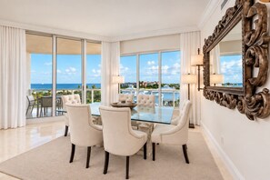 A spacious room with a glass table and four chairs, large windows with a view of the ocean, and a decorative mirror on the wall.
