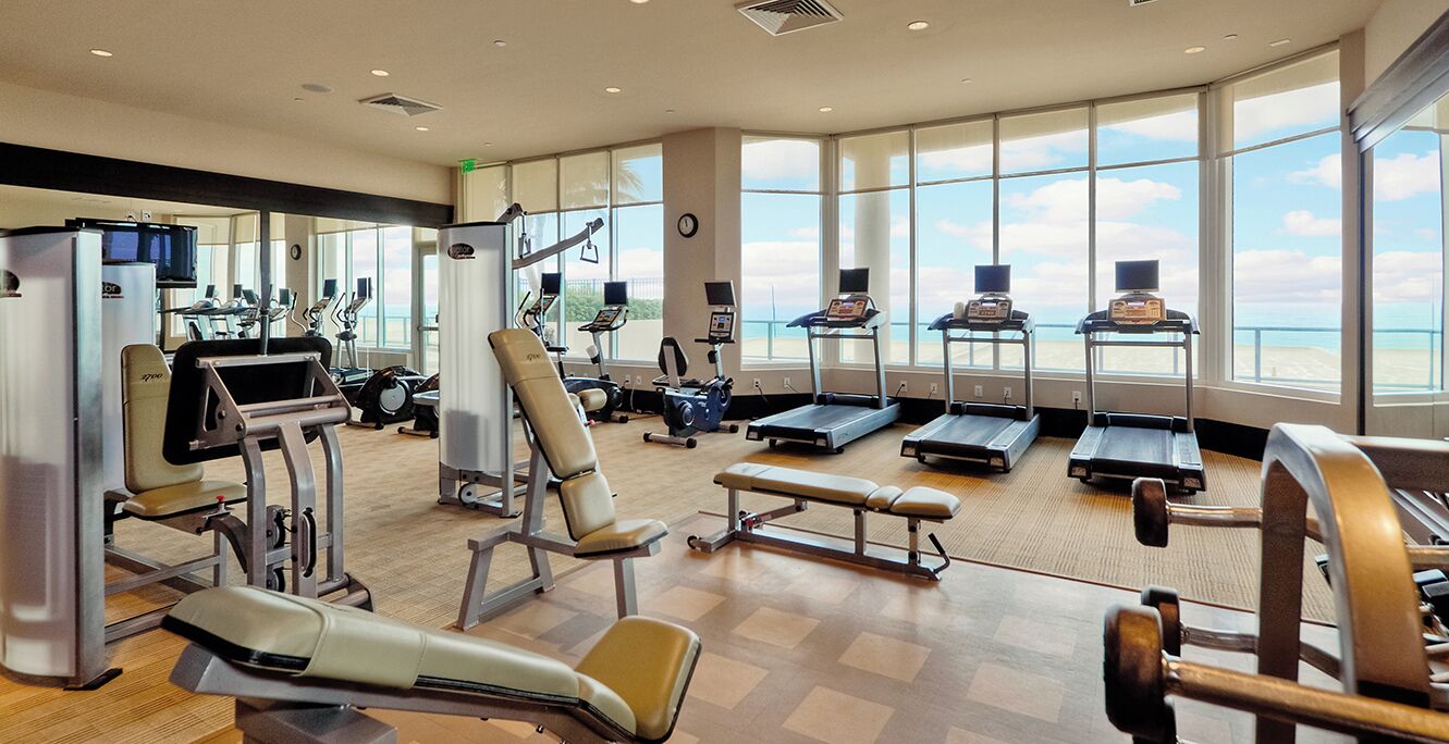 A spacious gym with various exercise equipment, large windows offering a view of the beach, and a modern interior design.