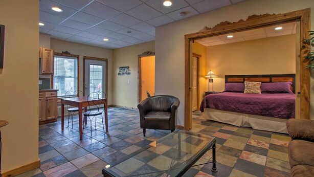 A hotel room with a checkered floor, a bed with a purple bedspread, a wooden headboard, a glass coffee table, and a leather armchair.