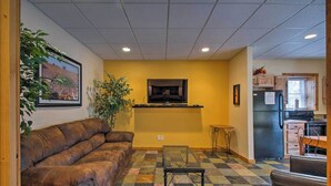 A compact living space with a sofa, a glass-top table, a TV mounted on the wall, and a kitchen area with cabinets and appliances.