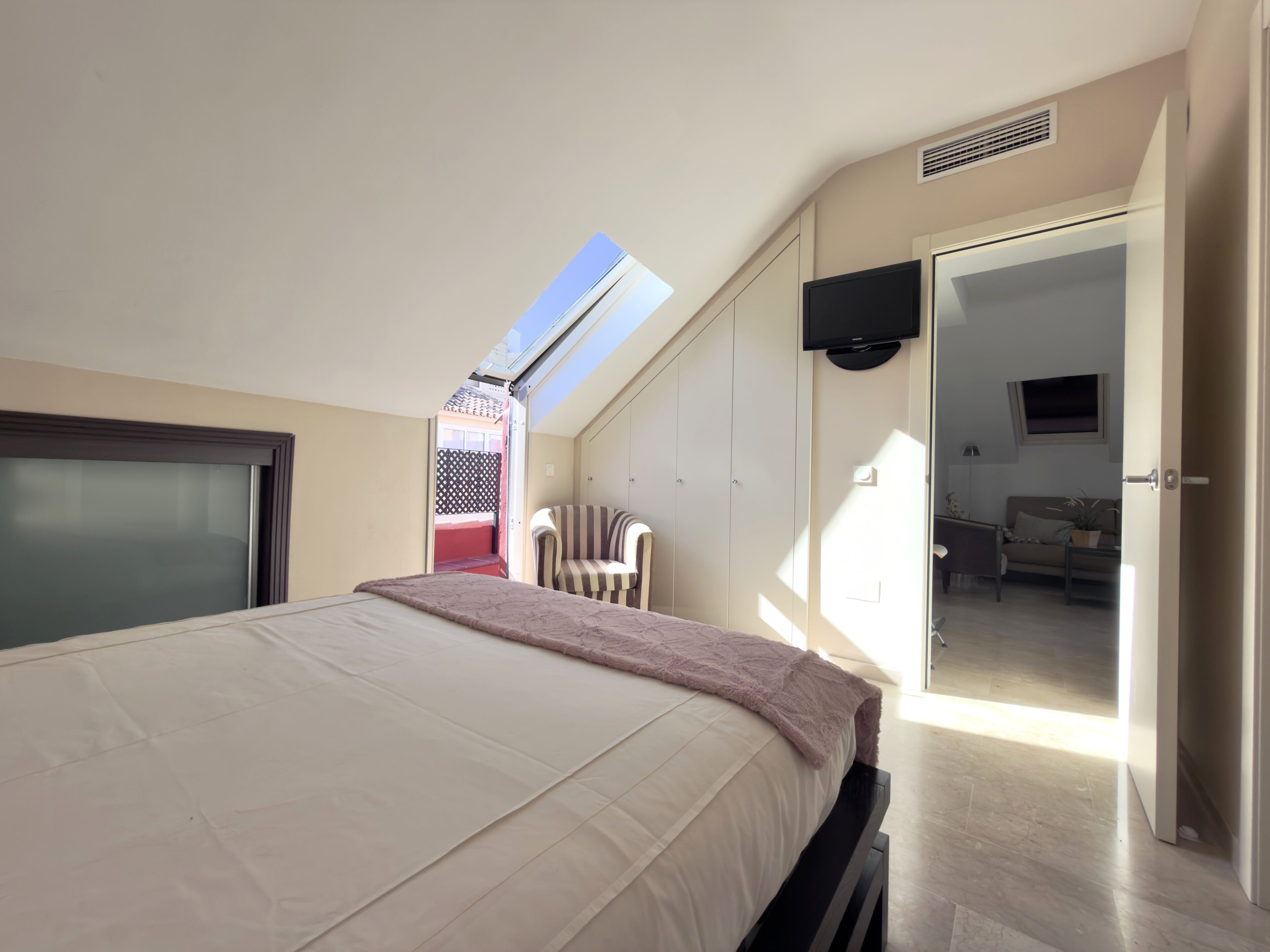 A bedroom with a bed, a TV mounted on the wall, a chair, and a door leading to another room.