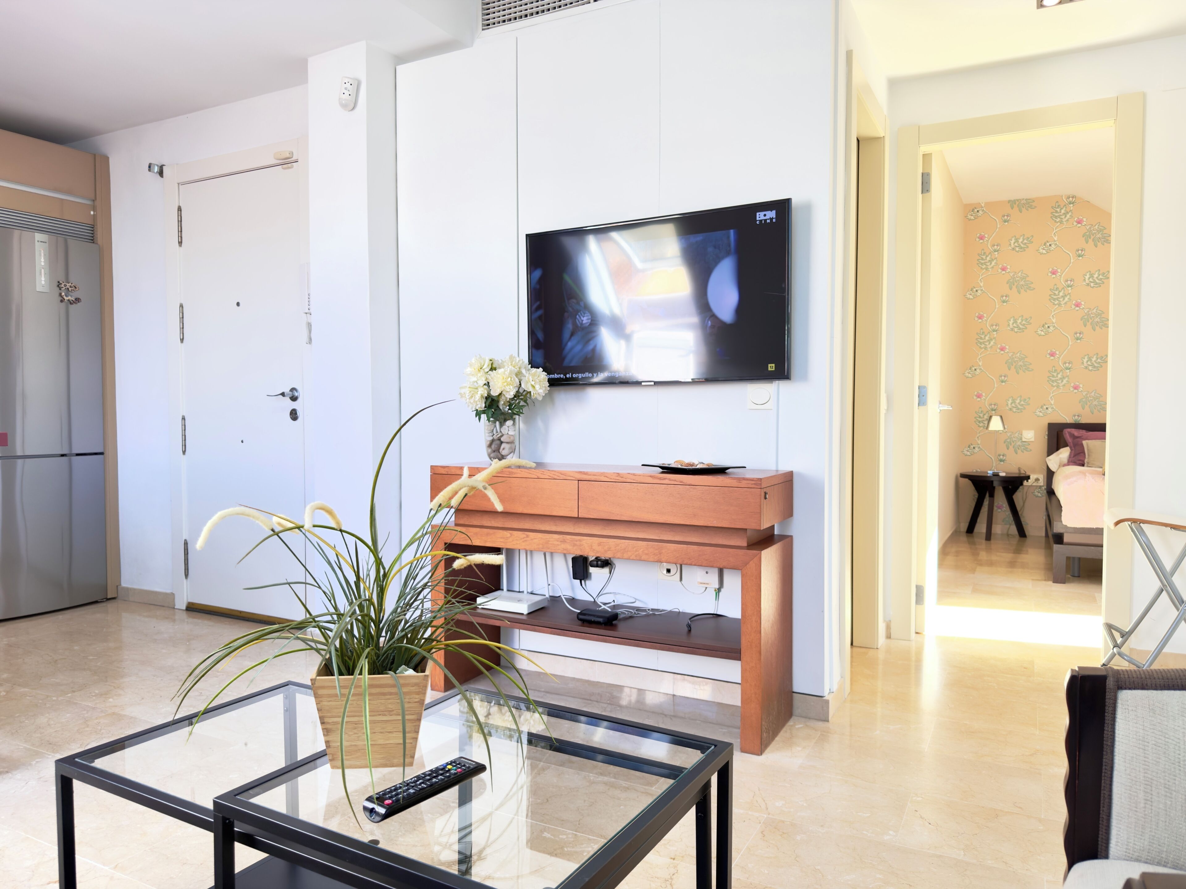 A modern living room with a flat-screen TV mounted on the wall, a wooden cabinet, a glass coffee table, and a potted plant.