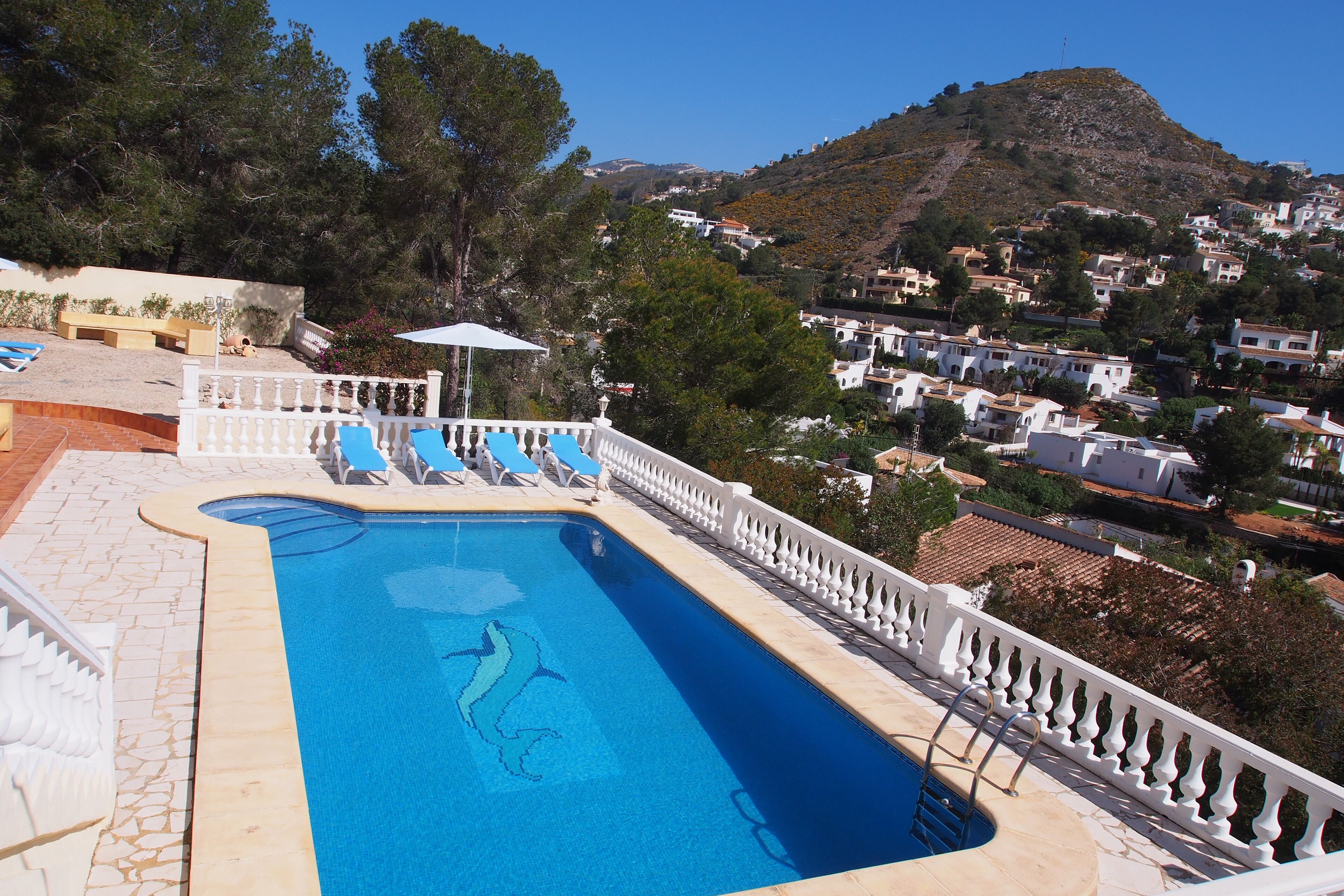 Une piscine décorée avec un motif de dauphin, entourée d’une balustrade blanche, et dont le décor est complété par une ville perchée sur une colline et des arbres.