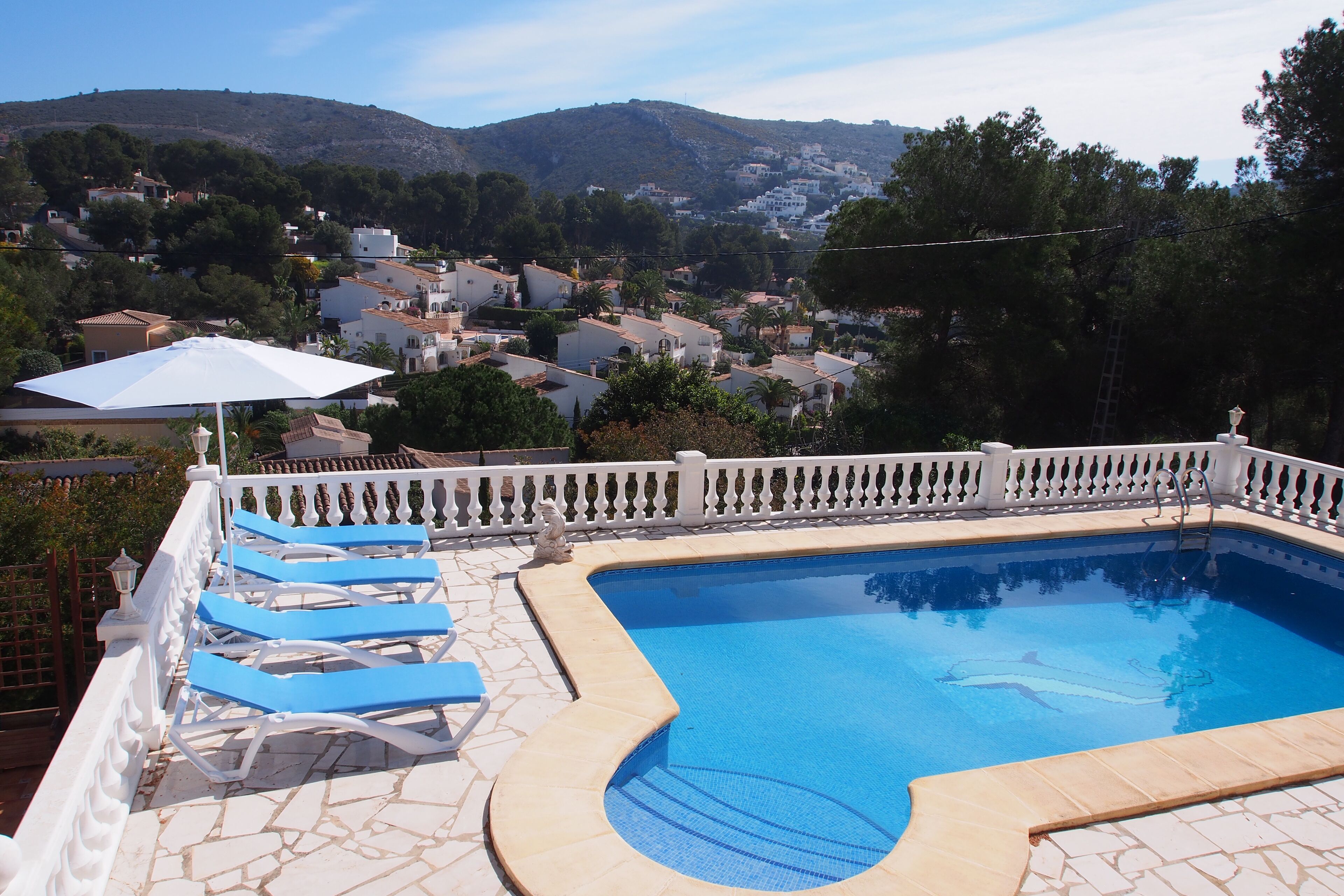 A swimming pool with blue lounge chairs and a white umbrella, surrounded by a white fence and a stone patio.