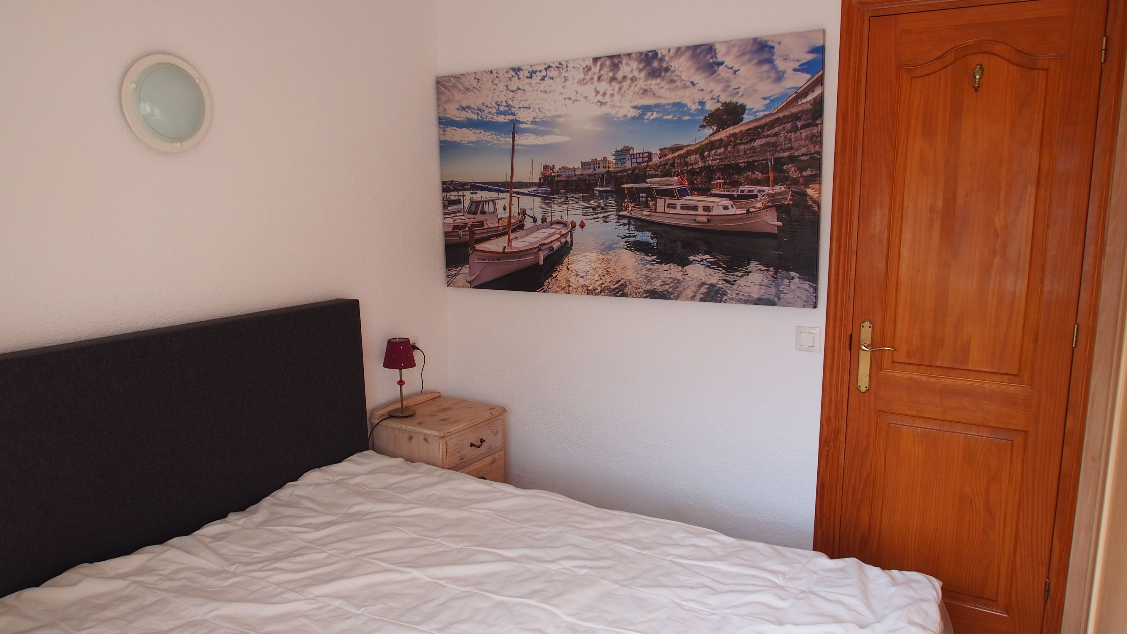 A bedroom with a bed, a wooden nightstand, a lamp, a door, and a wall-mounted picture of a harbor scene.