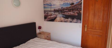 A bedroom with a bed, a wooden nightstand, a lamp, a door, and a wall-mounted picture of a harbor scene.