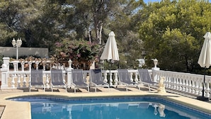 A poolside area with white chairs and umbrellas, surrounded by a white fence and trees.