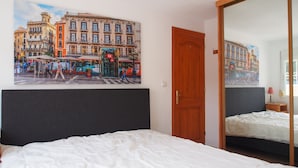 A hotel room with a large cityscape painting, a wooden door, a bed with white bedding, and a small bedside table with a lamp.
