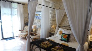 A four-poster bed with white curtains, a floral chair, a bedside table with a lamp, and a painting on the wall. - "Le Kiwi" - sleeps 2 self catering studio gite 10mins from Avignon! (Rognonas)