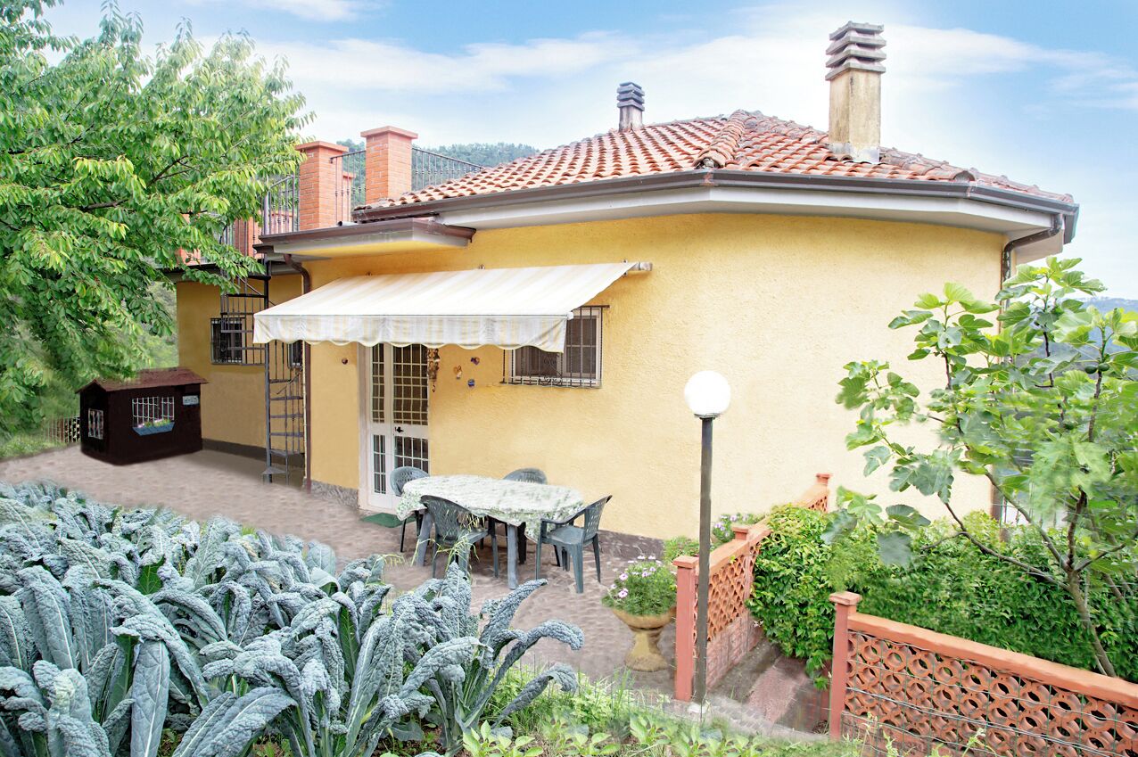 A yellow house with a tiled roof, a patio with a table and chairs, and a garden with large leafy plants.