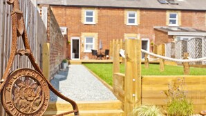 A rustic metal gate with a circular emblem, a wooden fence, and a brick house with a gravel path leading to the entrance. - Seaside Cottage with panoramic sea views. (Bournemouth)