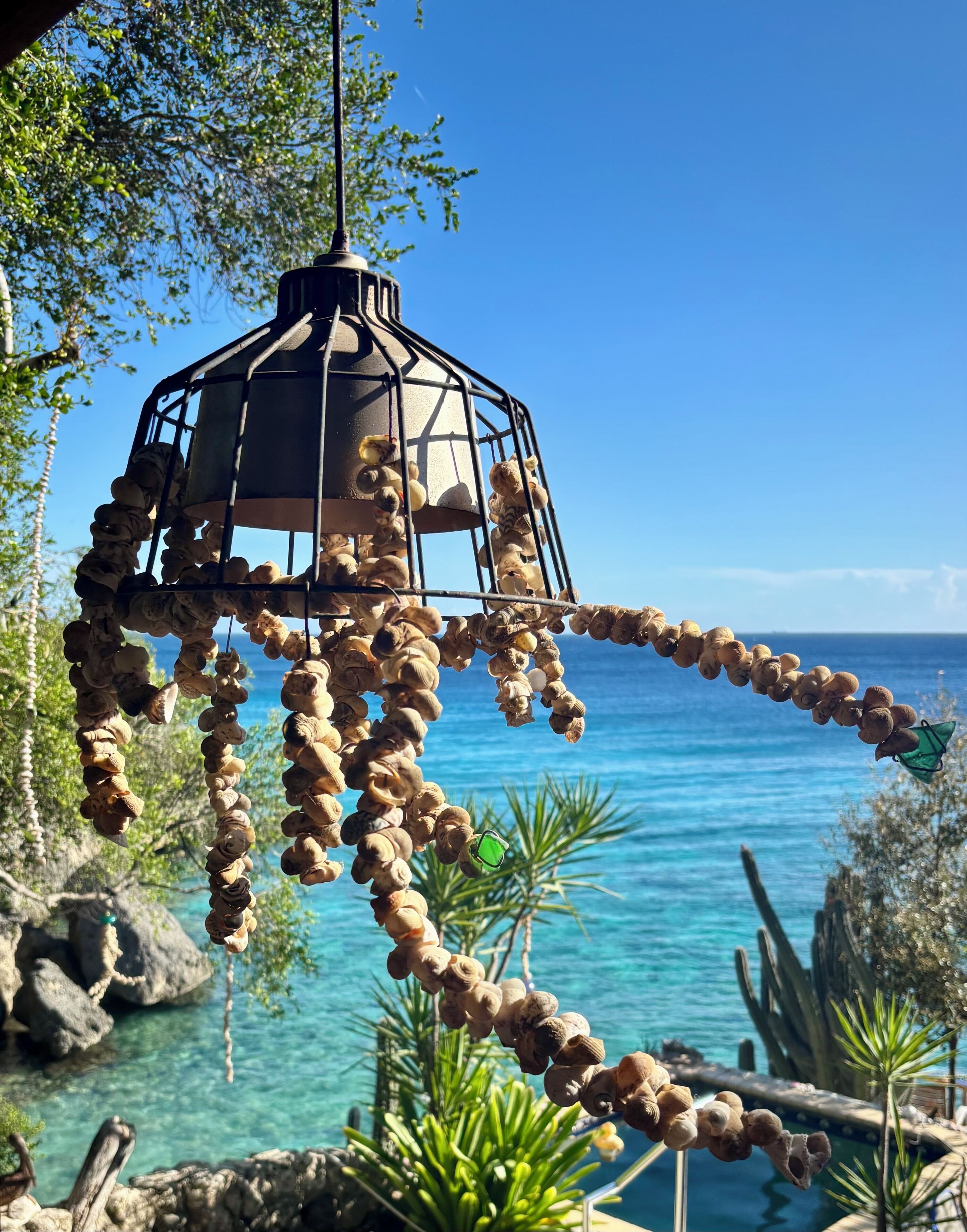 A hanging lantern with a spiral design, suspended outdoors with a clear view of the ocean and rocky terrain.