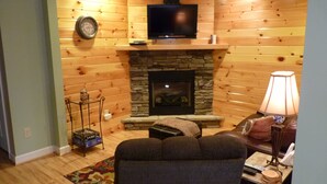 Un salon chaleureux avec des murs en bois, une cheminée en pierre, un téléviseur à écran plat, un canapé confortable et une petite table avec une lampe.
