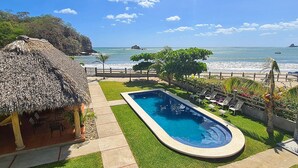 Una piscina con vista al océano, una construcción con techo de paja y un camino pavimentado.