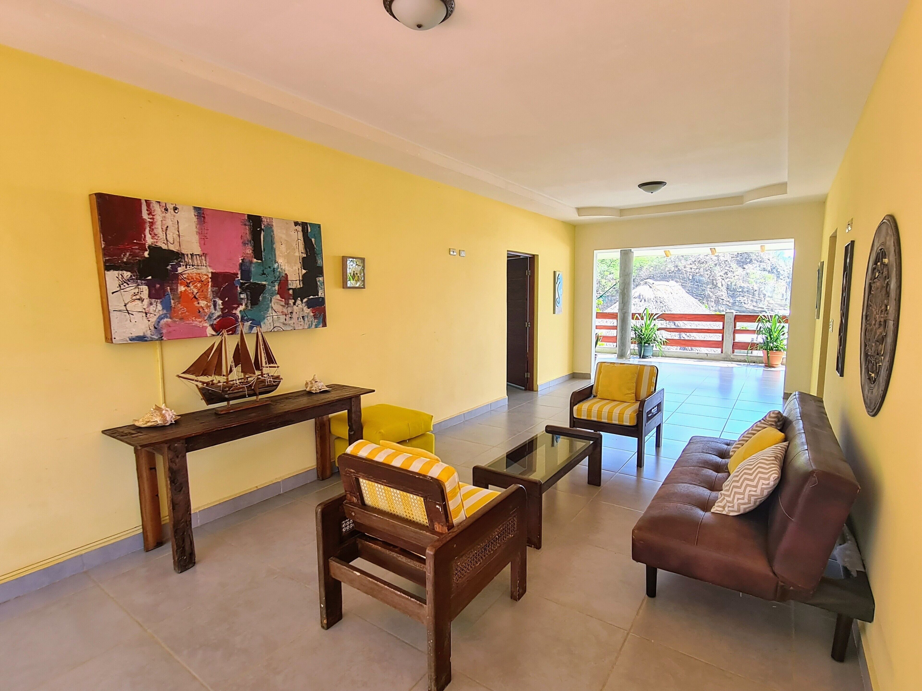 A living room with yellow walls, a wooden coffee table, a brown leather sofa, and a yellow armchair. There is a large abstract painting on the wall and a small ship model on a wooden table.