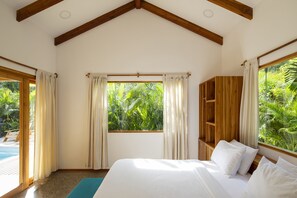 A bedroom with a large bed, wooden beams, a window with curtains, and a view of greenery.