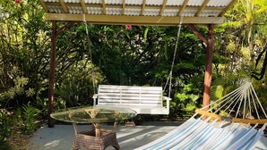 En hængekøje, en hvid bænk og et glasbord under et pergola i en have.