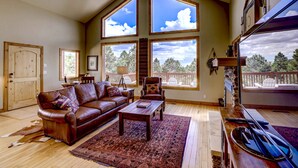A spacious living room with a large brown leather sofa, a wooden coffee table, and a red rug. There are large windows offering a view of the outdoors.