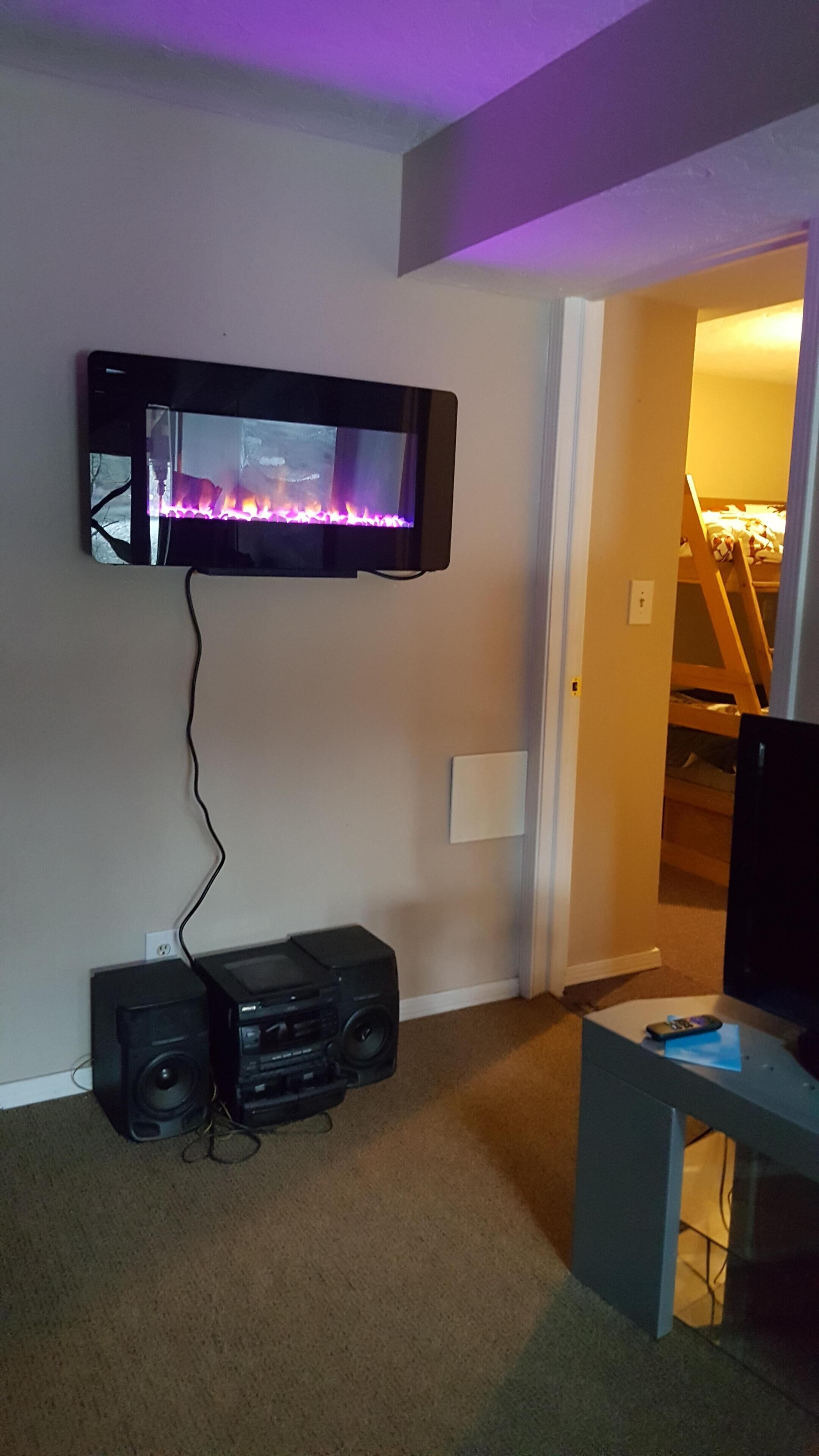 Habitación con televisor de pared mostrando un fuego, sistema de sonido y cama al fondo.